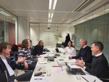 EEAC baord and secretarit members sit in a well-lit conference room around a table. Papers and laptops in front of them, all dicussing and smiling.